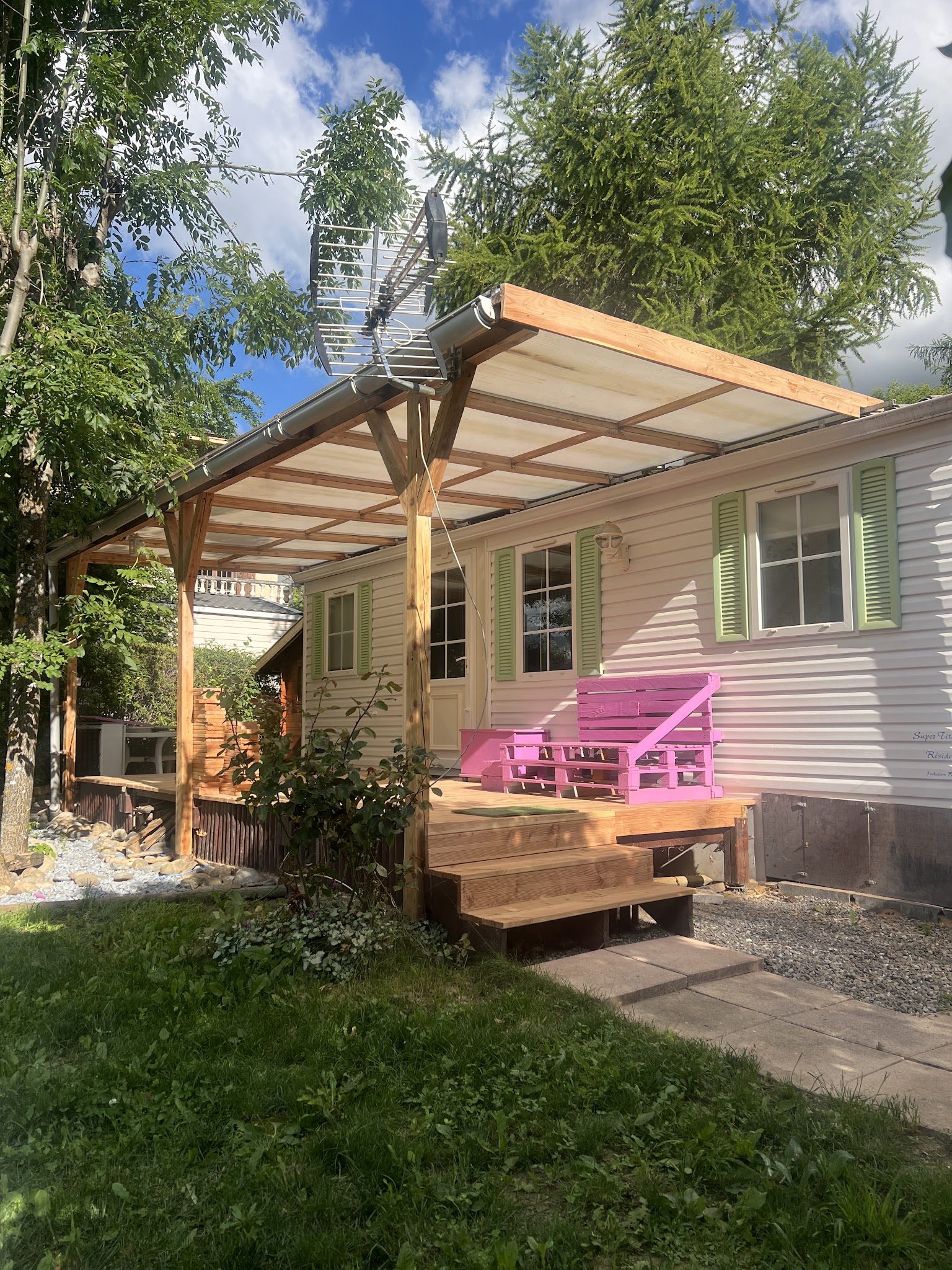Pergola en bois couverte de plaques translucides et terrasse en bois avec marches, devant une maison blanche aux volets verts
