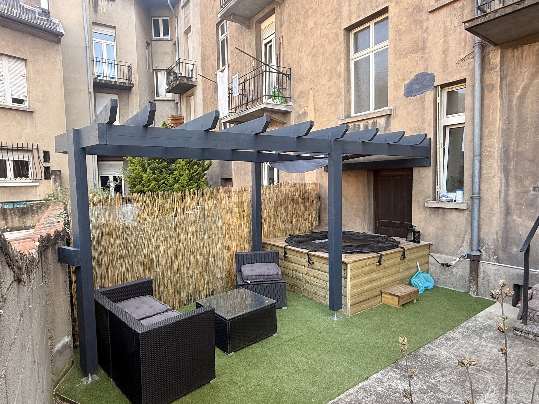 Pergola en bois peinte en gris anthracite dans une cour d'immeuble à Metz, salon de jardin et coffrage de spa en bois